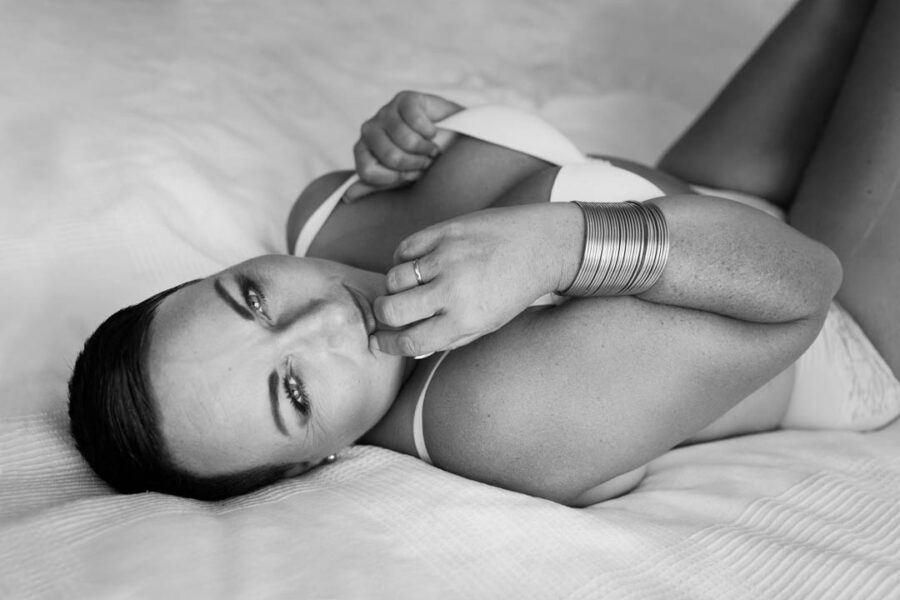 Sort-hvid boudoir af kvinde i hvidt lingerisæt, liggende sidelæns på seng og ser mod kamera