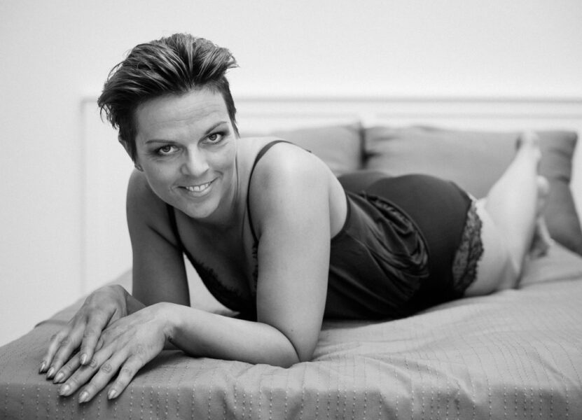 Sort-hvid boudoirfoto af kvinde i satinbody, liggende på maven på seng og smilende til kamera