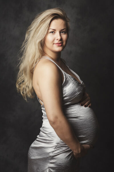 Studioportræt af gravid kvinde i sølvfarvet kjole, set i profil, der roligt holder om sin mave