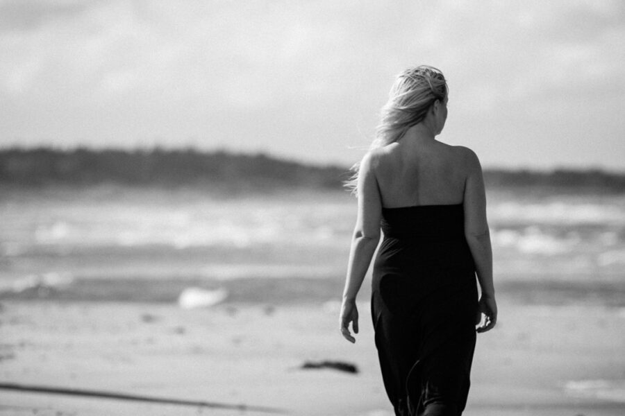 Stemningsfuld sort-hvid scene af kvinde i stropløs kjole, der går alene langs blæsende strand