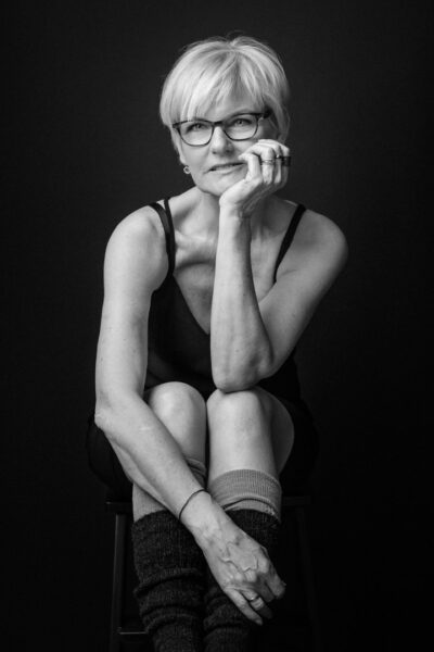 Sort-hvid studieportræt af moden kvinde på skammel i enkel dragt og benvarmere, rolig eftertænksom positur