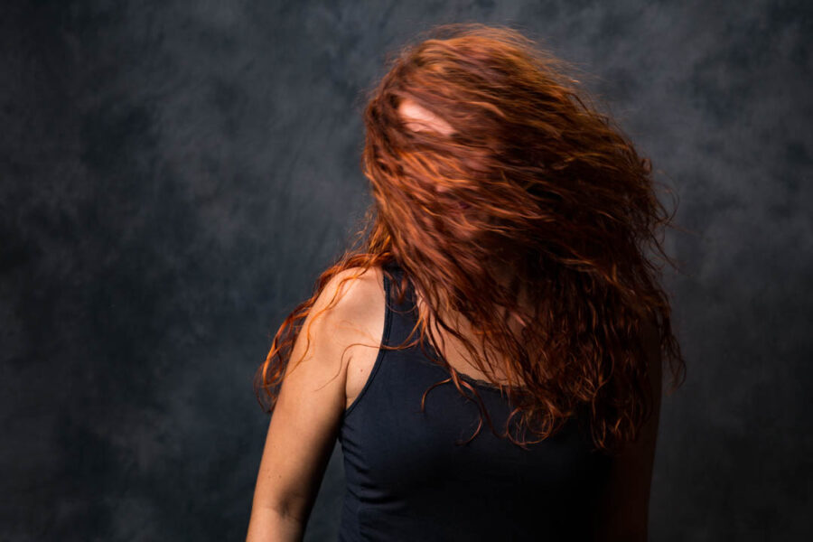 Farveportræt af kvinde i sort top, rødt krøllet hår svinger foran ansigtet mod mørk studiebaggrund